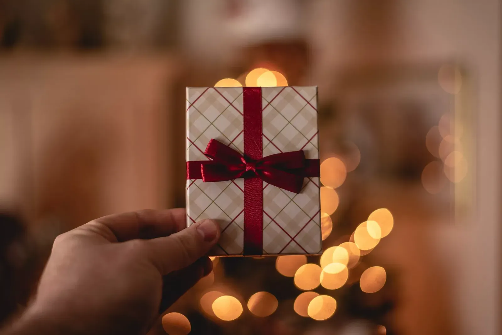 Mano que sostiene una cajita de regalo a cuadros con moño rojo frente a luces navideñas desenfocadas