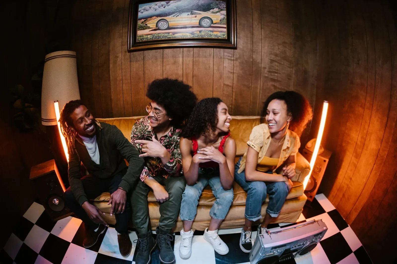 Cuatro jóvenes ríen sentados en un sillón amarillo en un living retro con paredes de madera y piso ajedrezado