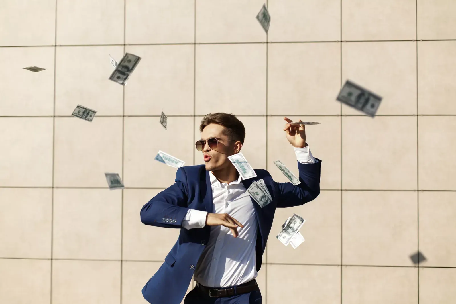 Hombre de traje azul y anteojos de sol lanza billetes de dólar al aire frente a una pared de baldosas claras