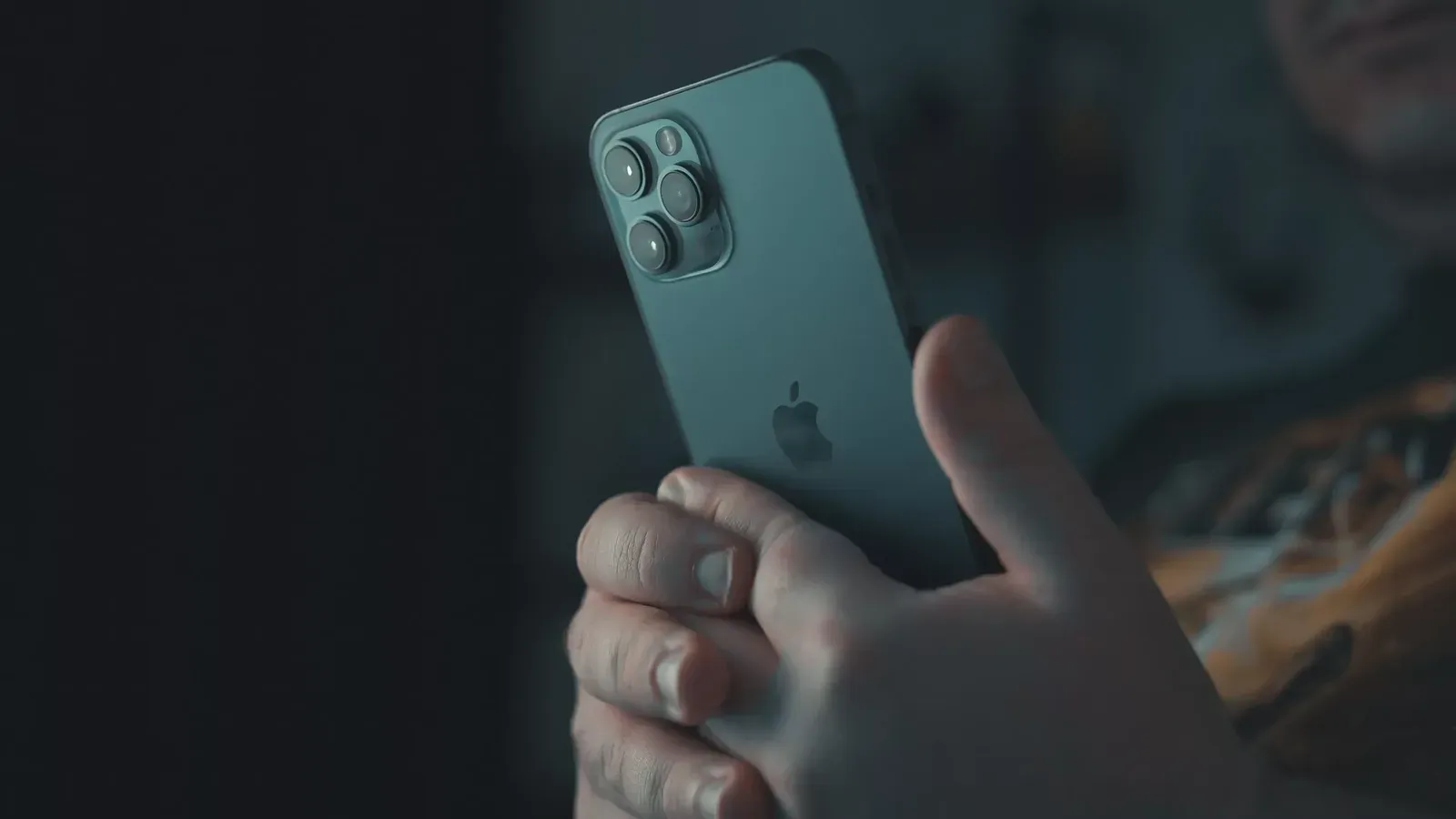 Primer plano de unas manos que sostienen un iPhone visto desde atrás en un ambiente en penumbra