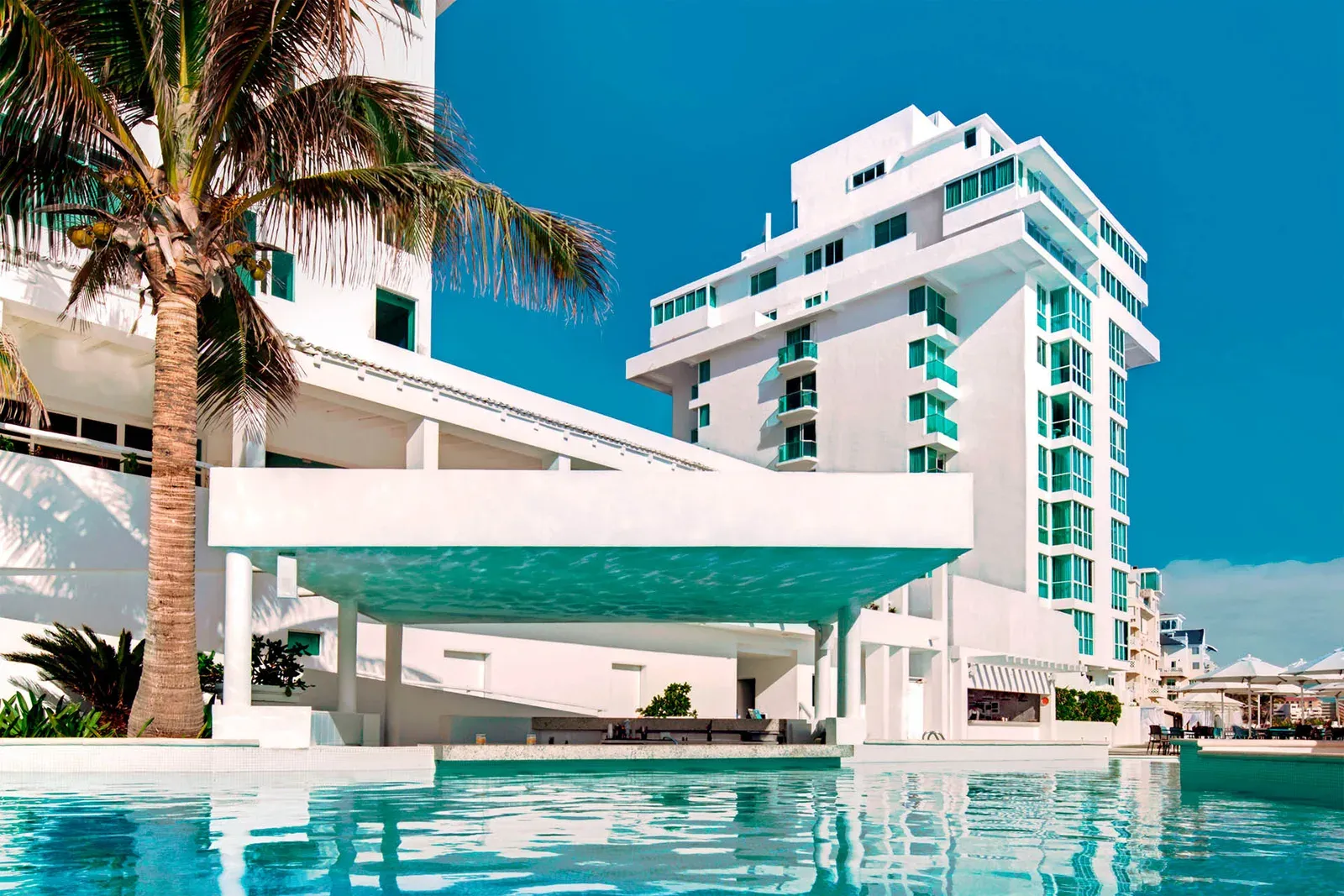 Hotel blanco de playa con pileta turquesa, palmera y cielo despejado