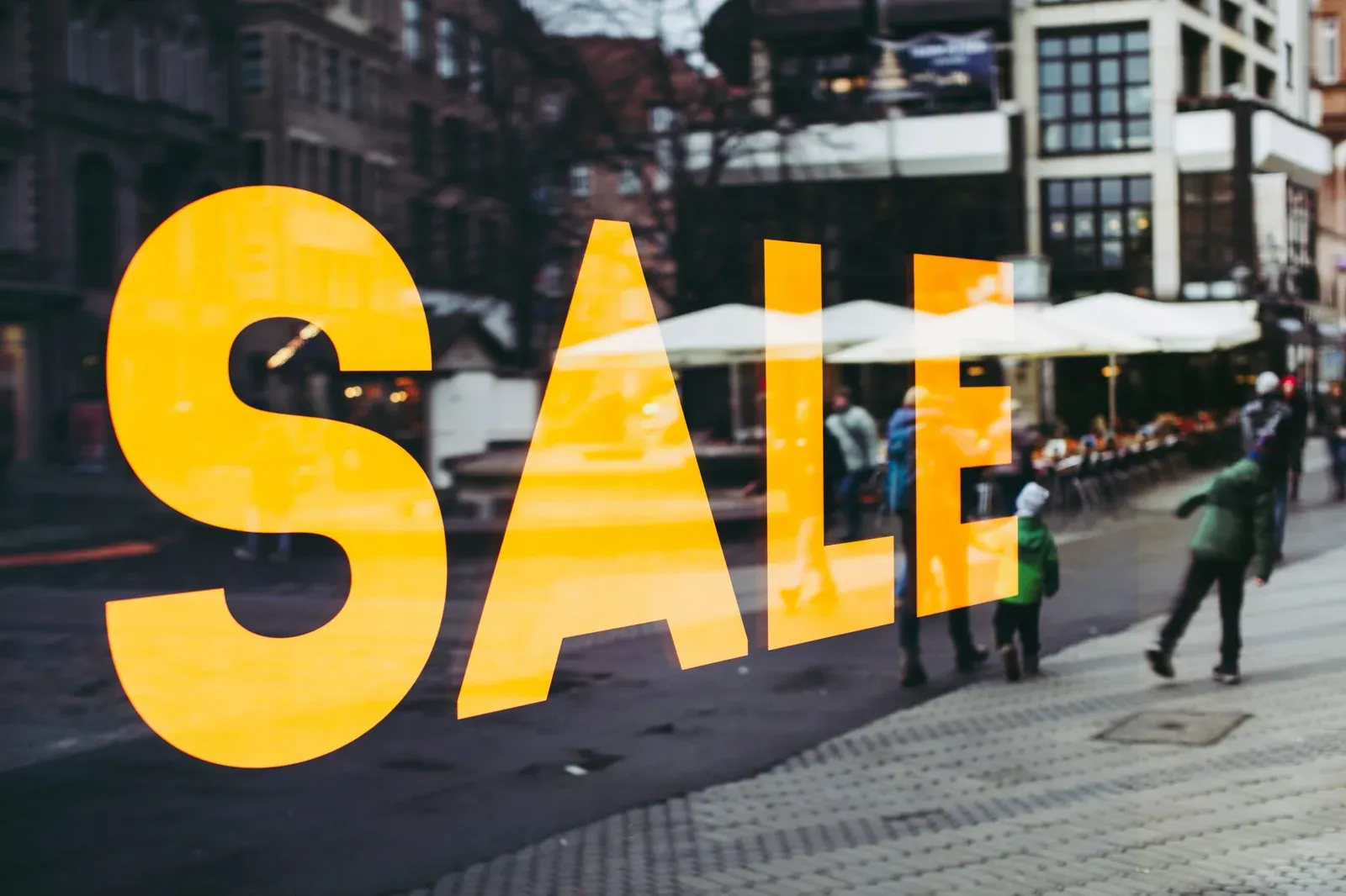 Cartel amarillo con la palabra SALE en una vidriera que refleja una calle peatonal con gente caminando