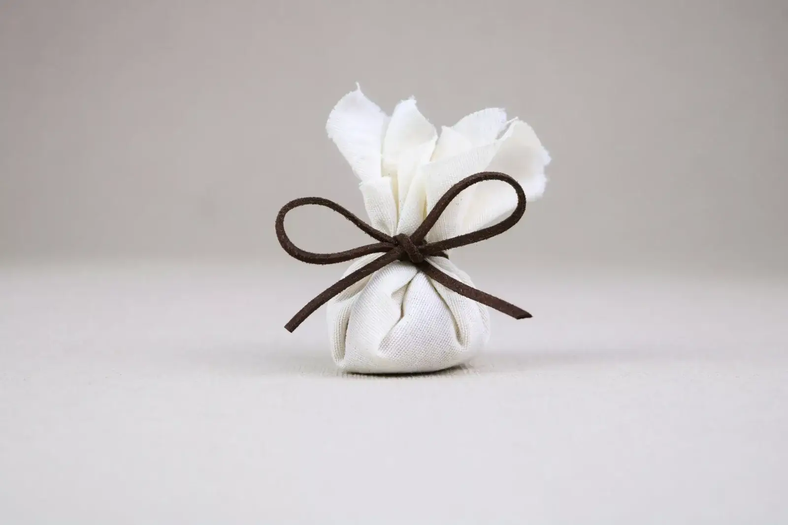 Pequeño regalo envuelto en tela blanca y atado con un moño marrón, sobre fondo gris