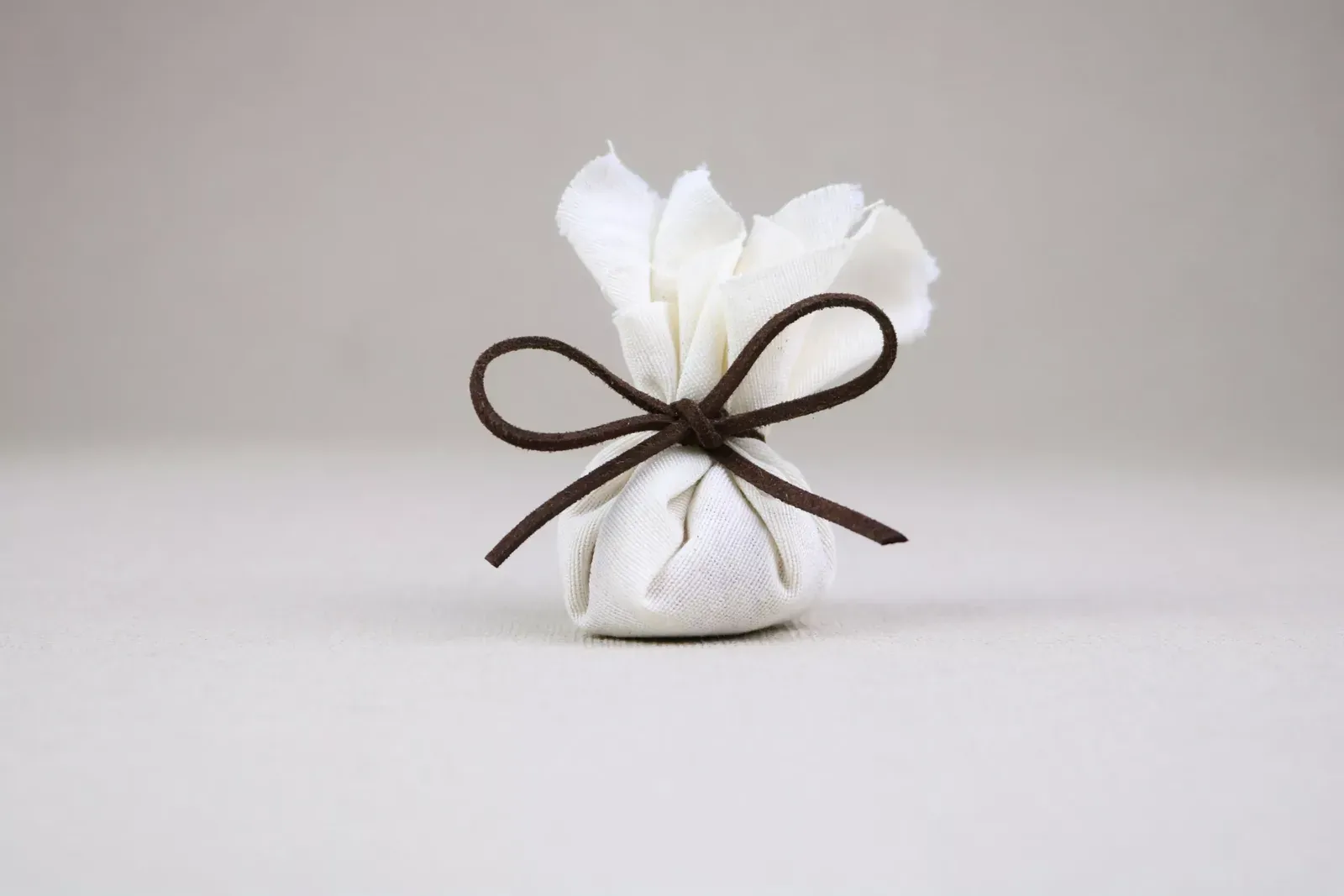 Pequeño regalo envuelto en tela blanca y atado con un moño marrón, sobre fondo gris