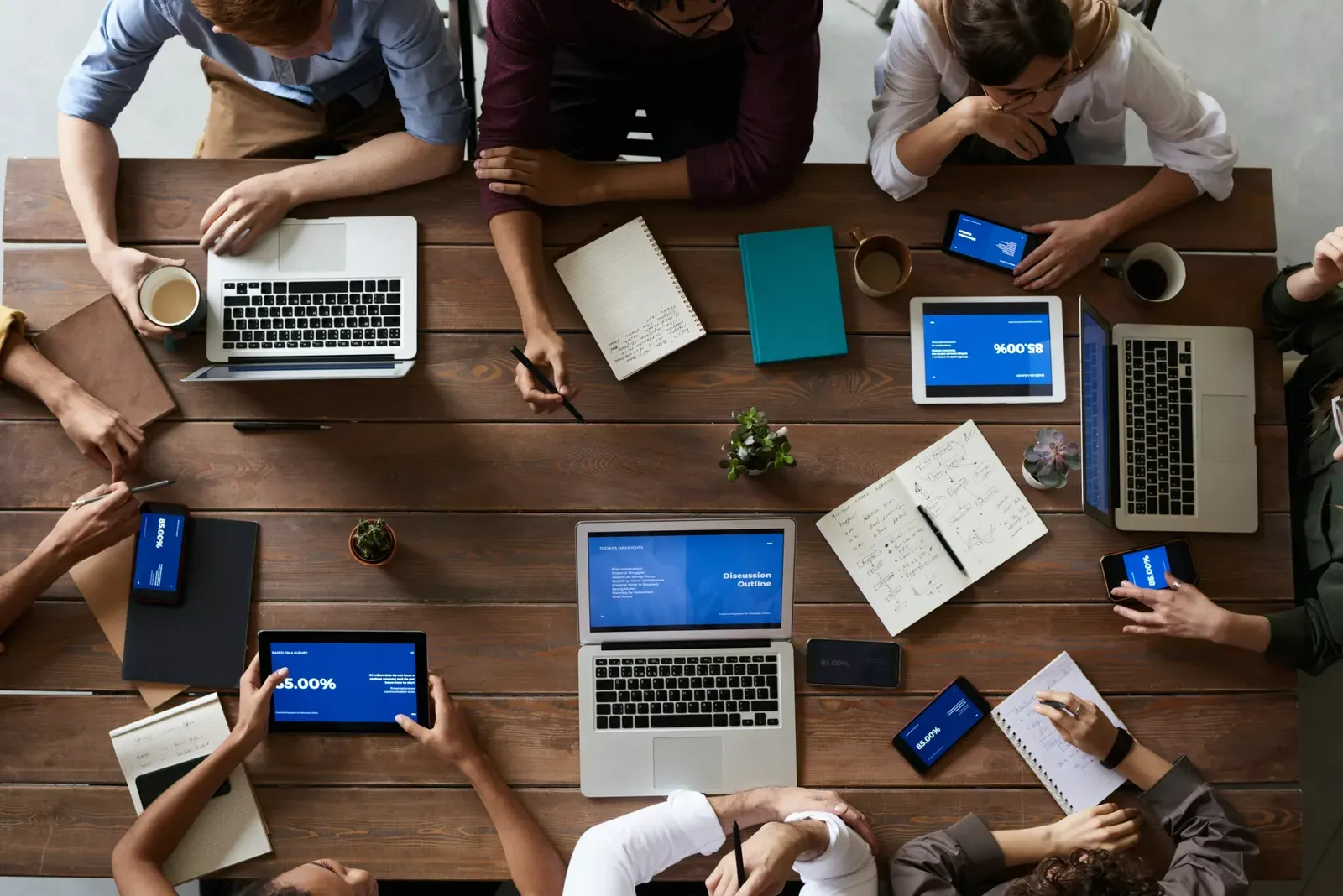 Vista cenital de un equipo reunido en una mesa de madera con laptops, tablets, celulares y cuadernos con notas