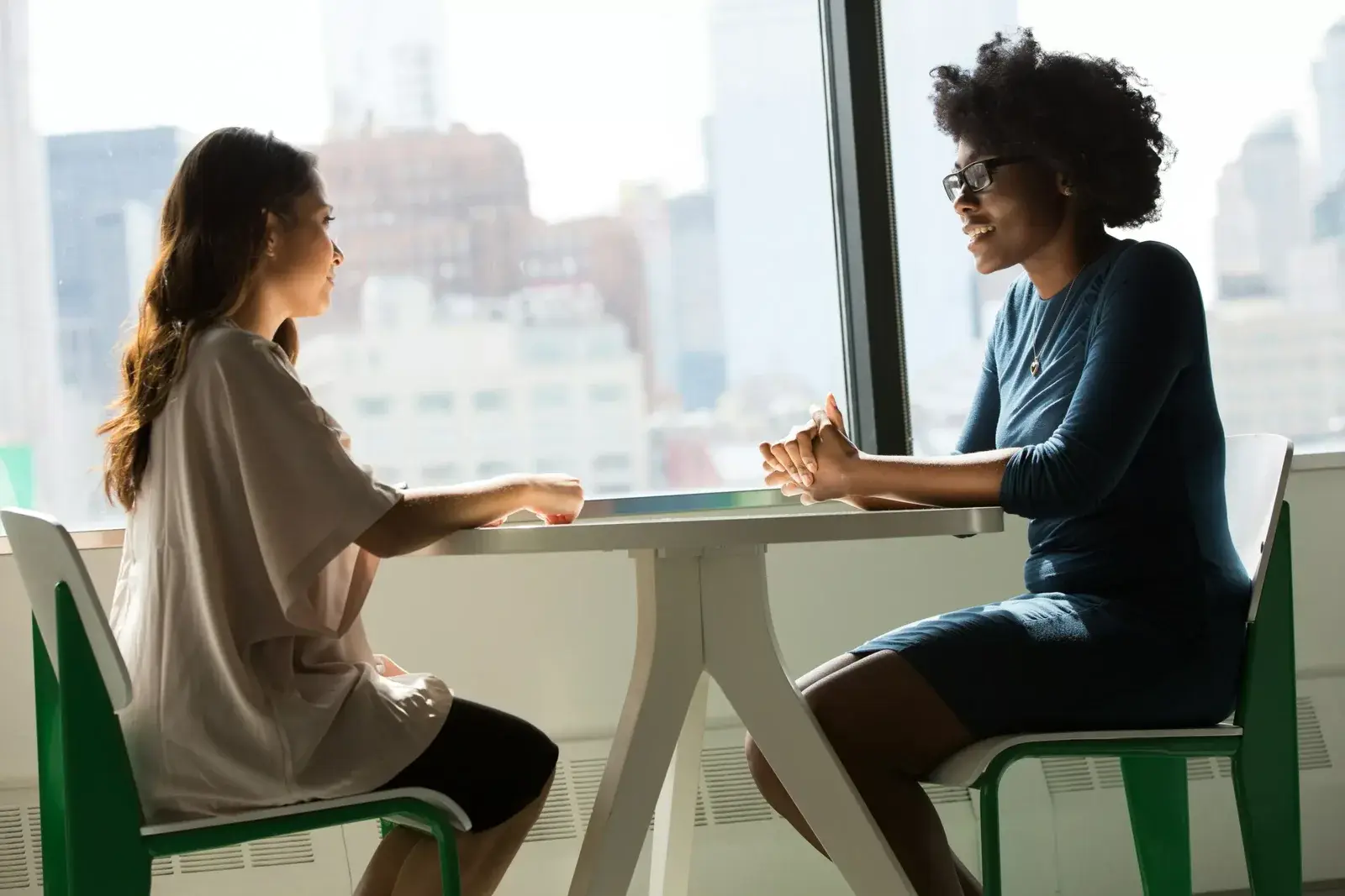 Dos mujeres conversan sentadas a una mesa junto a un ventanal con vista a la ciudad