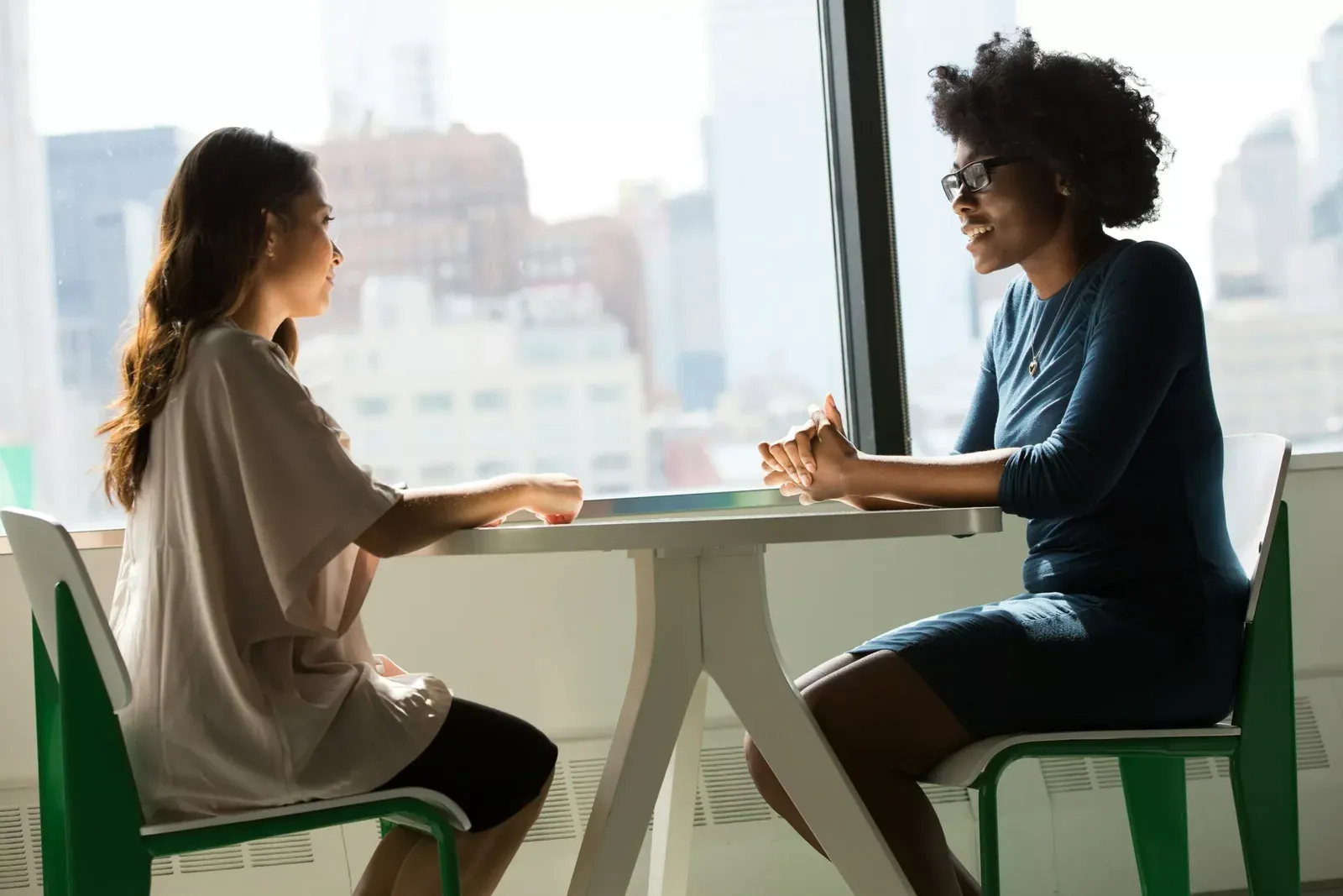 Dos mujeres conversan sentadas a una mesa junto a un ventanal con vista a la ciudad