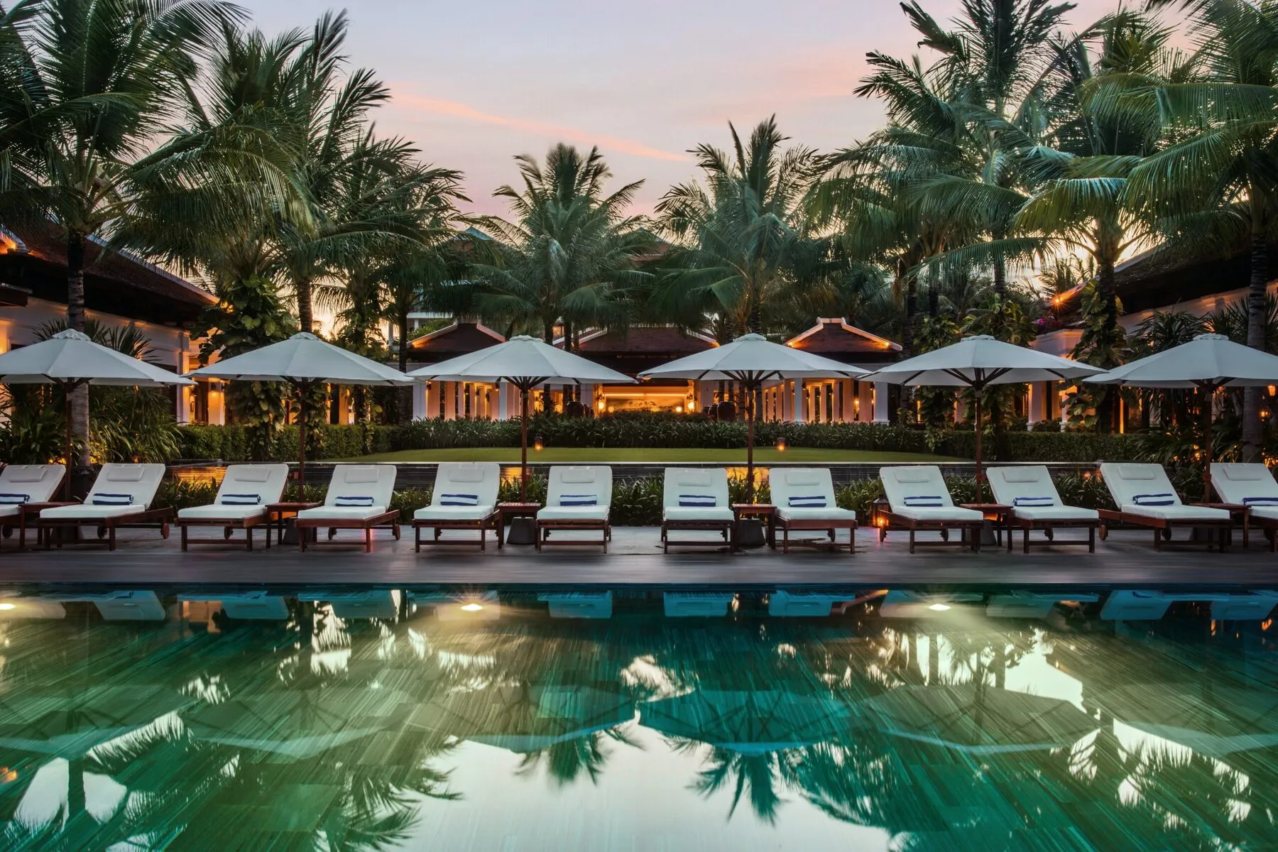 Pileta de un hotel al atardecer, rodeada de reposeras, sombrillas y palmeras