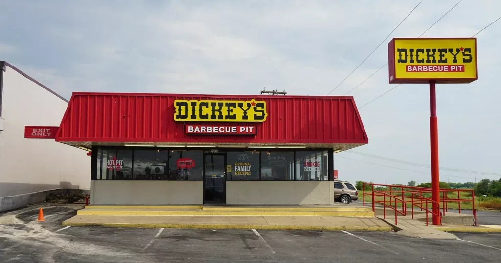 Fachada de un local de Dickey's Barbecue Pit con techo rojo, cartel amarillo y estacionamiento vacío