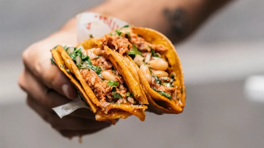 Mano que sostiene dos tacos de carne desmechada con cilantro, cebolla y porotos