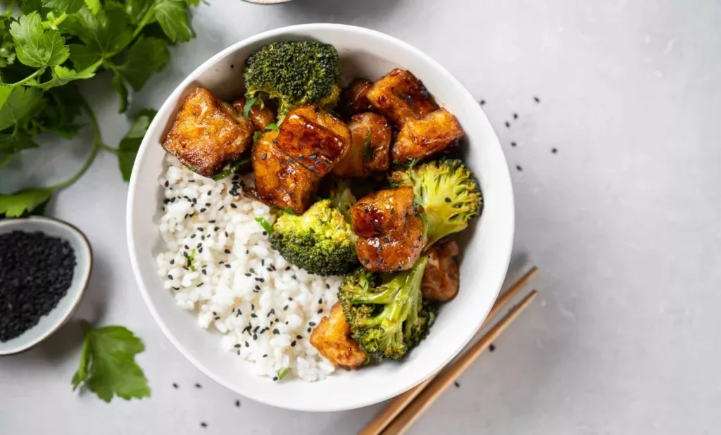 Bowl de arroz con tofu glaseado, brócoli y semillas de sésamo, visto desde arriba junto a unos palitos