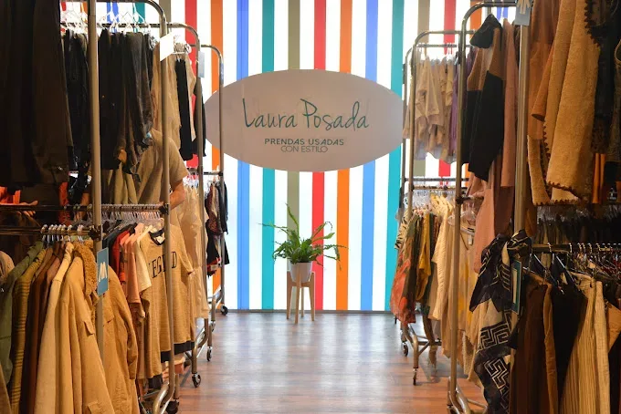 Interior del local de Laura Posada con percheros de ropa usada y el logo de la marca sobre una pared de rayas de colores