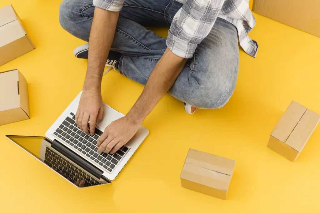 Persona sentada en el piso escribiendo en una laptop, rodeada de cajas de cartón sobre fondo amarillo